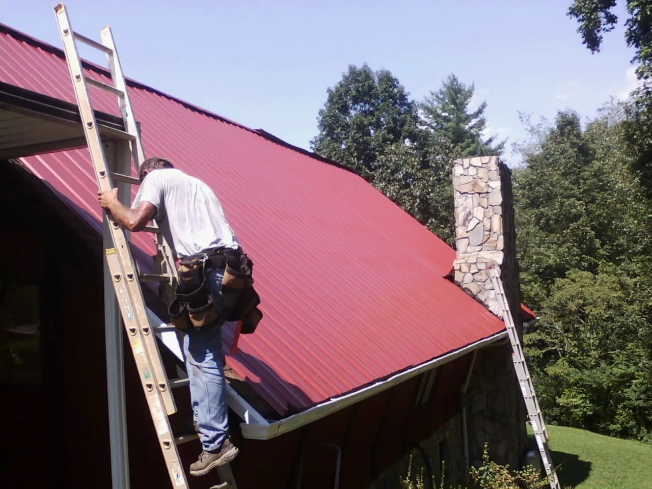 Roofer on a ladder beside a red metal roof and stone chimney — Soffit & Fascia Repair by Roofing Brothers in Woodinville